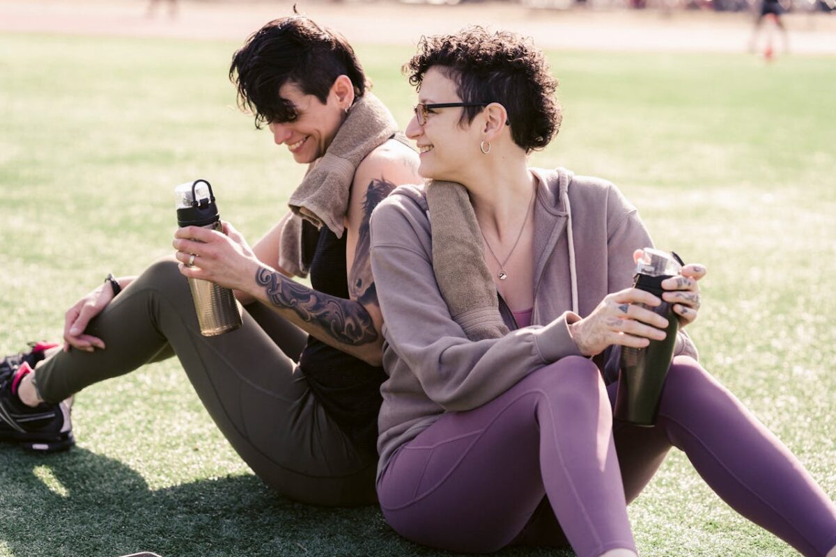 Couple of thirsty athletic girlfriends talking and smiling while drinking water and sitting on grass on stadium in back lit in daylight